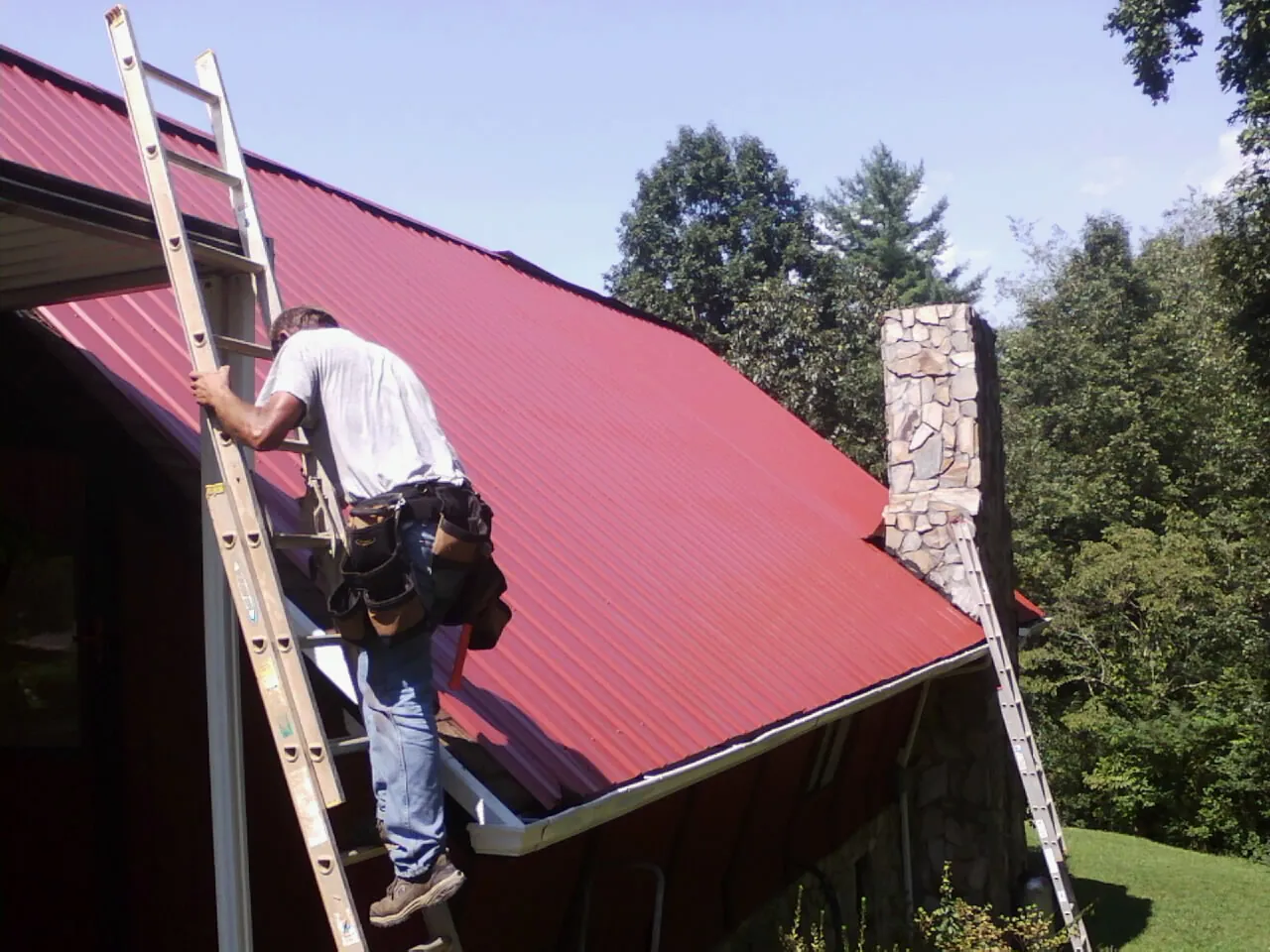 Roofer on a ladder beside a red metal roof and stone chimney — Soffit & Fascia Repair by Roofing Brothers in Charleston Afb